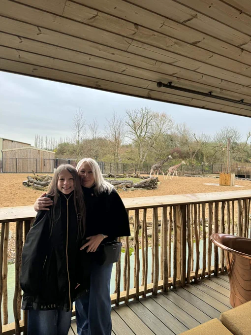 Adult woman hugging a smiling girl on a wooden viewing platform overlooking giraffes in a zoo enclosure.