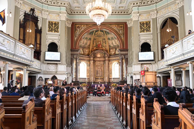 A large, ornate church interior with a high ceiling, chandeliers, and detailed artwork on the walls and ceiling. The church features a grand organ and large stained glass windows. A gathering of people is seated in the pews facing the altar.