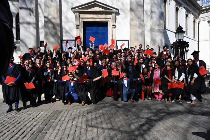 A large group of graduates in caps and gowns stand outside a building with a blue door, holding red diplomas and smiling. The scene is celebratory, with shadows of trees visible on the building facade.