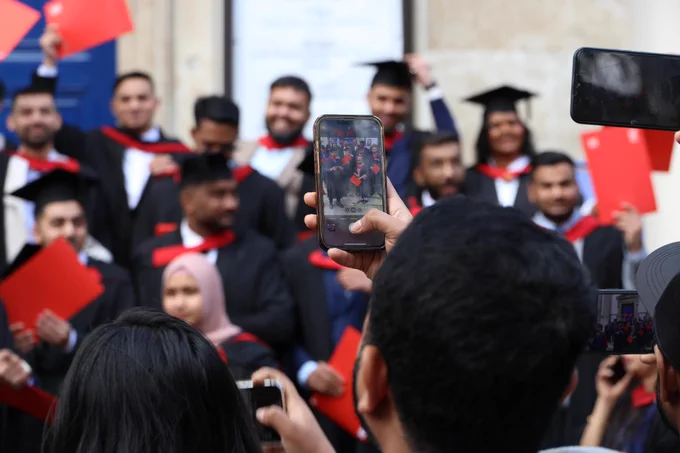 A group of people in graduation attire is posing for a photo. Some hold red diplomas. A person in the foreground is taking a picture with a smartphone. The setting appears to be an outdoor event.