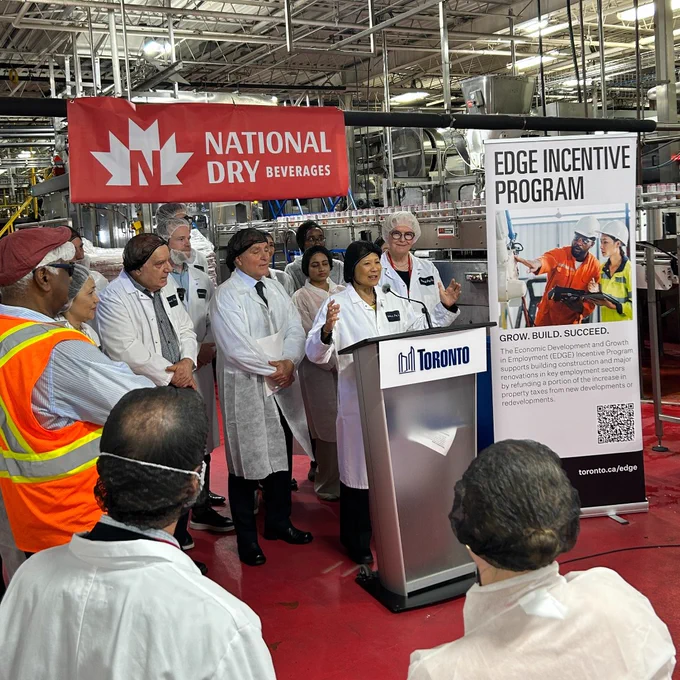 Mayor Olivia Chow speaks at a podium labeled Toronto inside beverage manufacturer National Dry with staff in lab coats and safety gear around her.