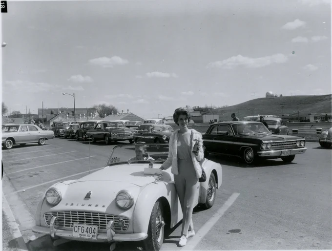 Black and white photograph from the WSU Libraries archives showing a person standing next to a convertible car in a parking lot. The car has a license plate reading “CFG 404” and the word “Pullman” on the front. Several mid-20th-century cars are parked in the background, and a grassy hill with buildings and utility poles is visible under a partly cloudy sky.