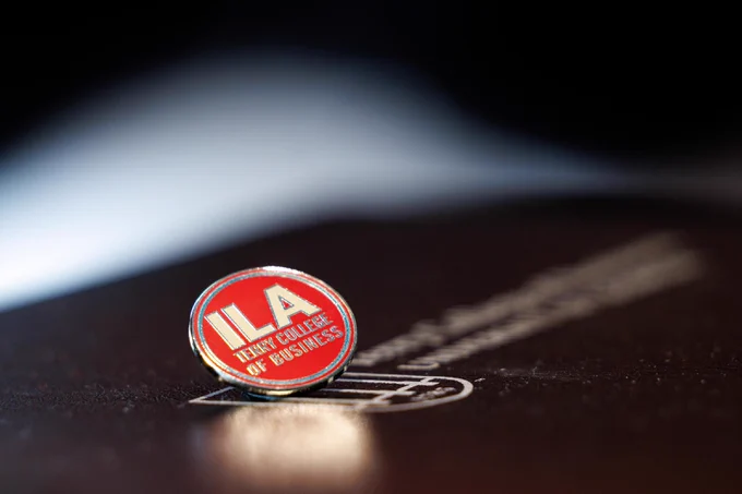 A circular pin displaying "ILA Terry College of Business" rests on a black Terry College of Business folder with soft lighting highlighting its shiny finish in front of a dark background.
