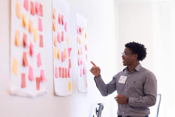 A student  gestures towards three large sheets of paper filled with colorful sticky notes on a wall.