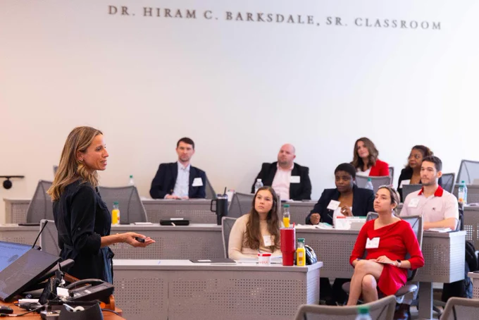 A woman stands at the front of a classroom speaking to seated individuals. The attendees are seated at desks with laptops and notepads. The setting is a formal presentation or lecture.