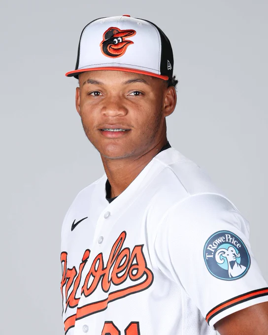 Luis De León looks at the camera and smiles. He is wearing a white Orioles jersey and a tri-color cap.