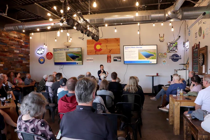 Group of people sitting in a brewery facing the front of the room where a woman is speaking into a microphone and two TVs are displaying information about Highlands Ranch Water's surface water storage reservoirs behind her.