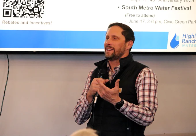 Man speaking into a microphone with a TV displaying information behind him.