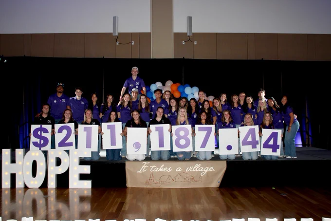 The Relay for Life Team stands on a stage smiling with signs that read $211,187.44. There is a banner with the words "It Takes A Village"
