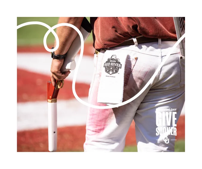 A person stands on a football field holding a rifle and a towel with a visible "Red River Rivalry" logo on it. They are dressed in white pants and a crimson shirt. The background features a blurred red and white design on the field.

GiveSooner and University of Oklahoma logos are featured as well.