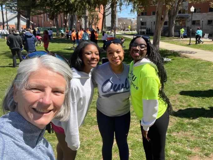 A group of four individuals is standing outdoors on a grassy area during a sunny day. They are smiling for the camera. The setting appears to be a park or a campus with other people and buildings in the background.