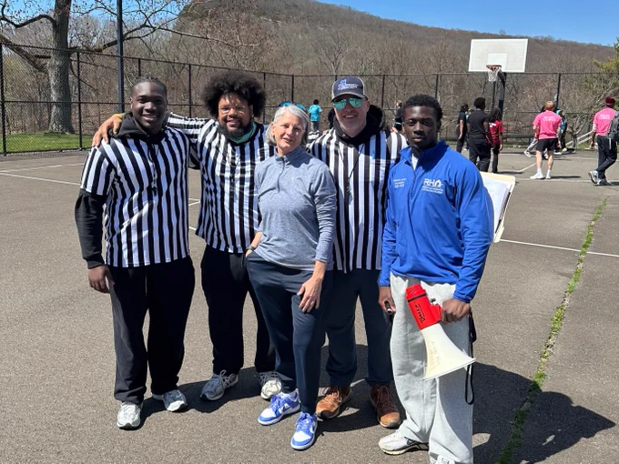 A group of five people stand together on a basketball court outdoors. Three are wearing black-and-white striped shirts, one is in casual sportswear, and another is holding a red-and-white megaphone. A basketball hoop and other individuals are visible in the background.