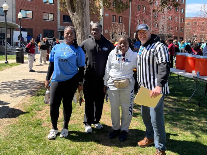 A group of people smiling and standing outdoors on a grassy area at an event. They are casually dressed, one person wearing a striped shirt is holding a folder. A few leafless trees and a red-brick building are in the background.