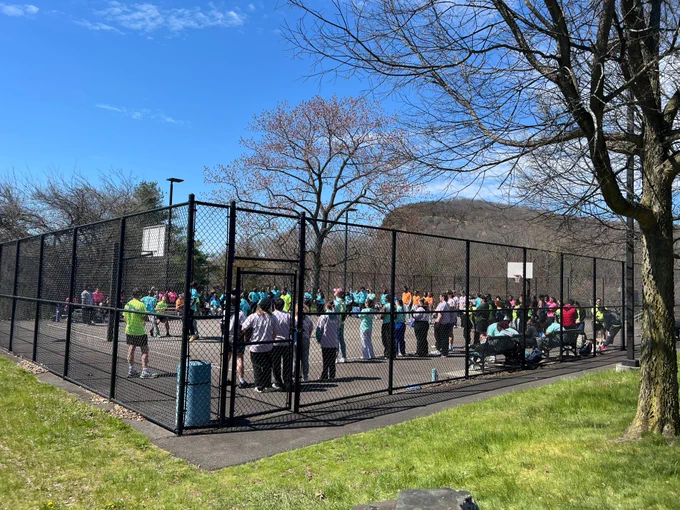 A group of people gathered inside a fenced basketball court, wearing colorful shirts and helmets, possibly participating in an event. A few trees and hills are visible in the background under a clear blue sky.