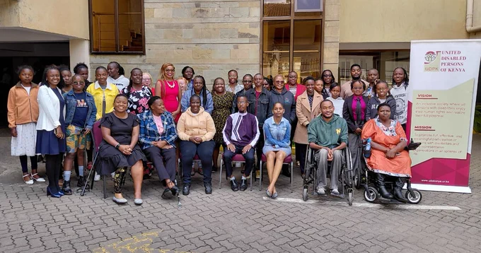 A large group of participants pose for a group photo outside a building, representing diverse persons with disabilities and allies. Some individuals are seated in front while others stand behind, including participants using wheelchairs and crutches. A UDPK banner is displayed beside the group, highlighting the organization’s vision and mission. The setting reflects a collaborative and inclusive workshop or consultation bringing together disability rights advocates.