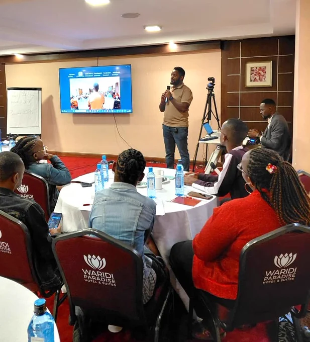 A man stands at the front of a conference room speaking into a microphone while participants sit around round tables listening attentively. A screen behind him displays a virtual meeting, showing hybrid participation. A camera on a tripod records the session, and another facilitator sits nearby with a laptop. Attendees, seated in branded conference chairs, have notebooks and water bottles on the tables, indicating an interactive workshop or training environment.