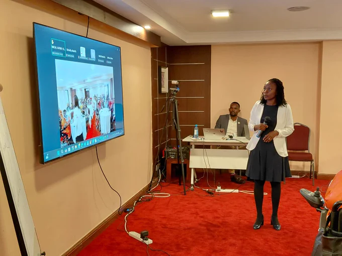 A woman stands at the front of a conference room holding a microphone, presenting to participants while facing a large screen displaying a virtual meeting. A camera on a tripod records the session, and a facilitator sits at a desk behind her with a laptop. The setup shows a hybrid workshop environment, combining in-person and online participation.