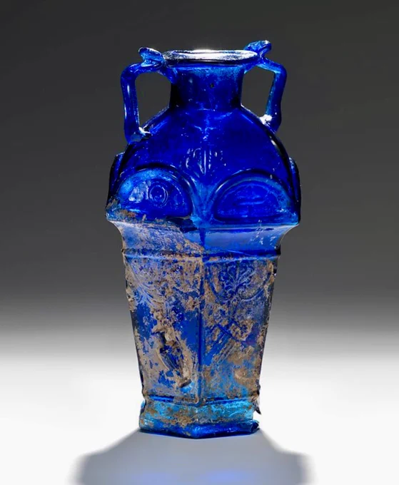 The Met photo shows a mould-blown translucent dark blue hexagonal-shaped flask (amphoriskos) signed in Greek with the name of the glassmaker ‘Ennion’ (not seen in image). Displayed against a graduated dark to light grey background.

The flask has a short cyclindrical neck, hexagonal sloping shoulders curving inwards towards the hexagonal straight-sided body which tapers downwards towards the base.  It has two thin handles attached at the top of the shoulders and top of the rim.

The mould blown glass has relief decoration associated with the god Dionysus and his retinue on the shoulders and five of the six body panels; double flutes, pan pipes, wine cup, wine jug, and vine leaves and grapes. On the sixth panel is the Greek inscription in three lines of parallel text  ENNIWN/EΠWH/CEN translated as ‘Ennion made me/it’.

The bottom of the flask is broken in places and the body has patches of creamy brown weathering with faint iridescence. Height: 14.3 cm, width 7.9 cm, depth: 7.2 cm