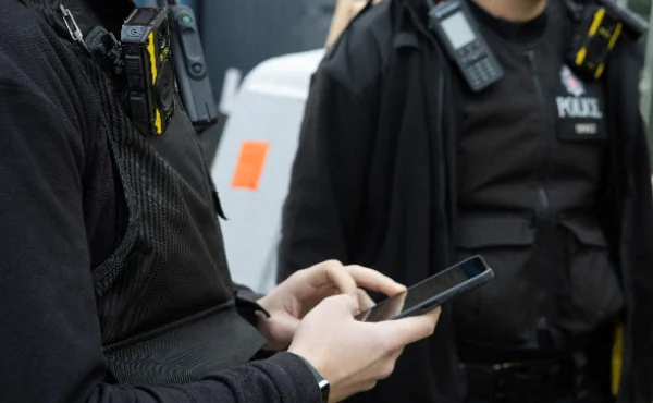 A photo of two police officers from the neck down, one of whom is typing into a mobile phone.