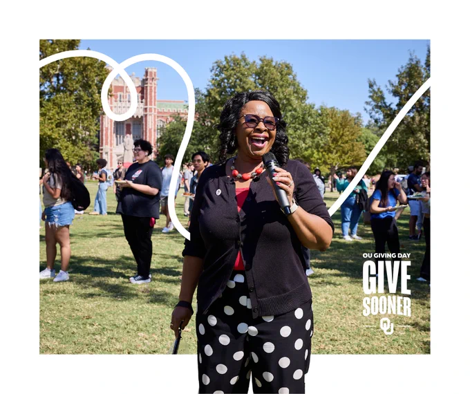 A person speaking into a microphone is standing outdoors on a grassy area. Trees and a large crowd are in the background. The University of Oklahoma's Bizzell Memorial Library can be seen in the distance. The image includes text: "OU Giving Day - Give Sooner.