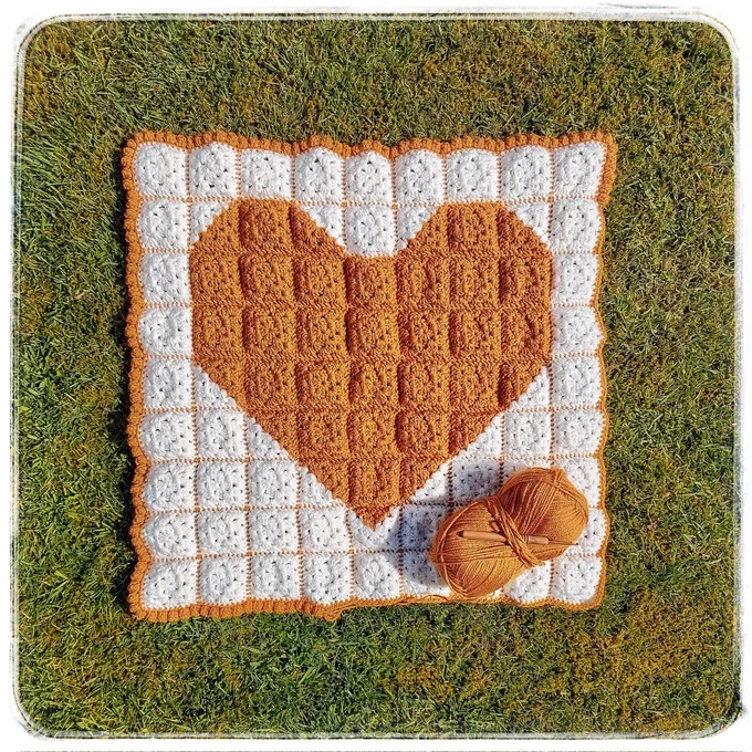 A crochet blanket in the making of a large yellow heart patchwork style on a cream background, with a bobble edging almost finished, the crochet hook & ball of working yarn sat on the corner, all spread out on mossy filled green grass.