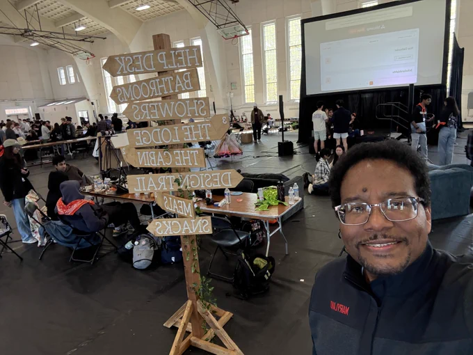 President Pines takes a selfie featuring a rustic wooden signpost with directions to various event areas like the "Help Desk" and "Design Trail."