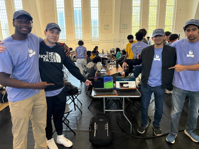 Four Terps wearing matching blue shirts pose together while pointing toward two open laptops on a table.