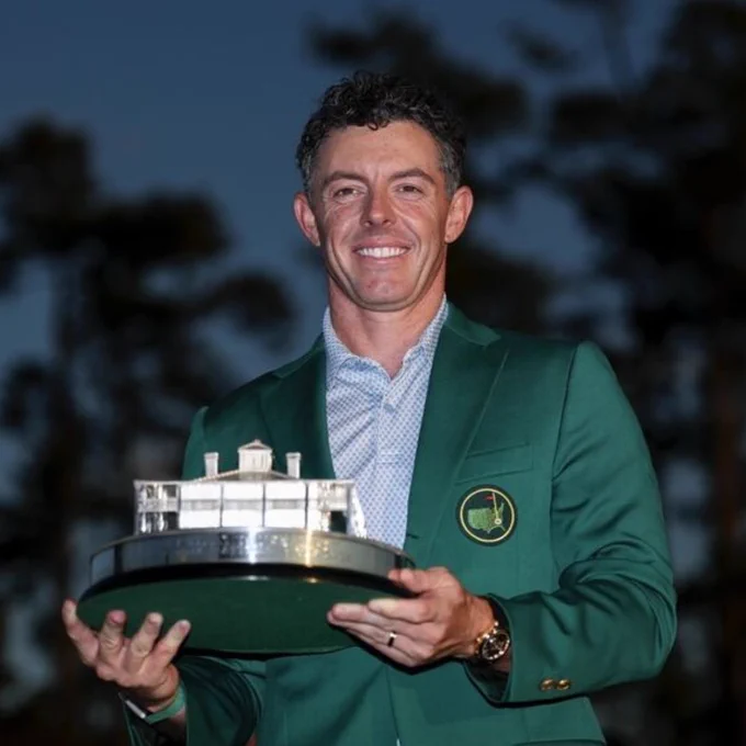 Golfer Rory McIlroy in a green Masters jacket holding the iconic Masters Tournament trophy at dusk outdoors.
