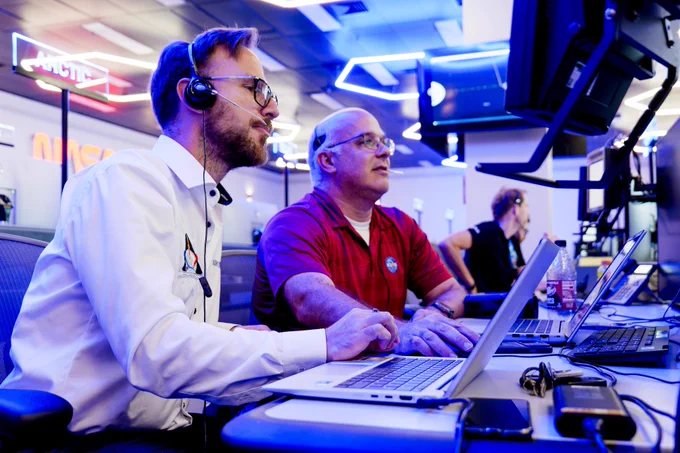 Two NASA employees wearing headsets work at laptops in a mission control room, focused on monitors displaying data.