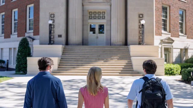 Three students stand with their backs to the camera facing the front steps and entrance of a large brick academic building with columns.