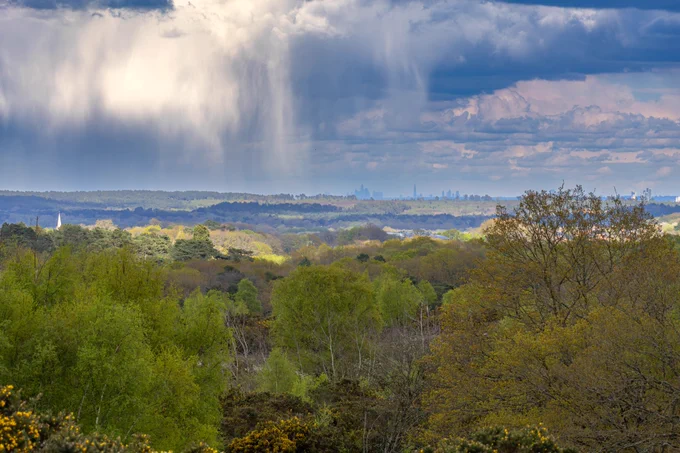 Landscape photograph of rain storm approaching Caesar's Camp Hill Fort with London just visible on horizon, Farnham, Surrey, UK, 16 April 2024.