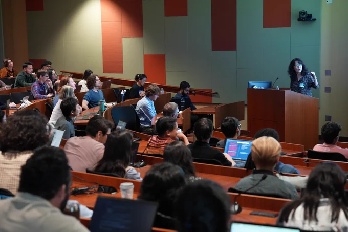 A lecture room with attentive students seated in rows facing a woman presenting at a podium. The room is well-lit, creating a focused atmosphere.