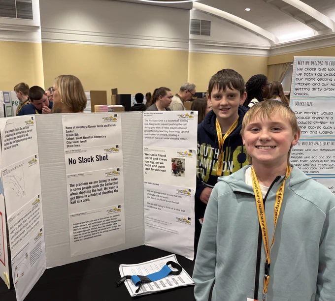 Two boys pose by a tri-fold board titled “No Slack Shot,” a basketball training aid, in a crowded exhibition hall.