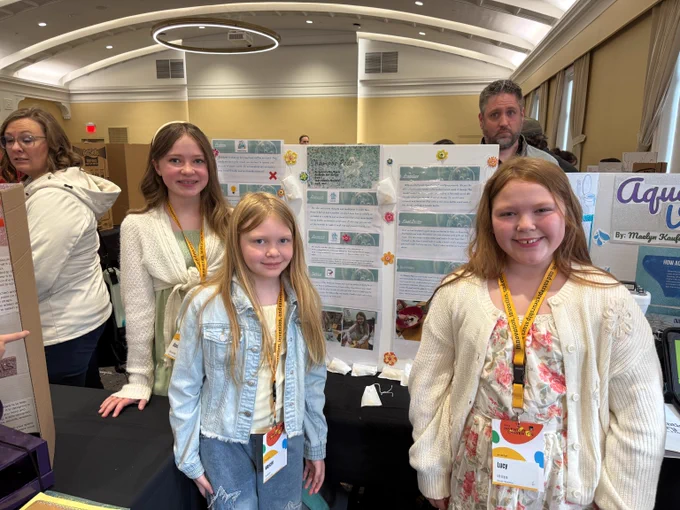 Three girls stand at a project display about shampoo pods at a busy invention fair, smiling in a bright conference room.