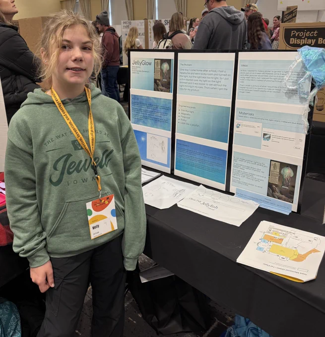Student named Molly stands beside her “JellyGlow” project board featuring a jellyfish-inspired light design at a fair booth.