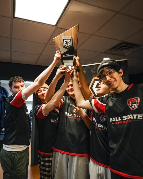 Five Ball State Esports players celebrate together, arms raised as they hold up a Collegiate Esports Valorant Champion trophy.
