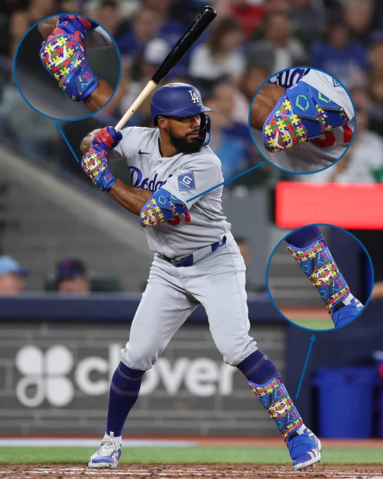 Teoscar Hernández stands in the batter’s box during an at-bat. Three zoomed-in images around him show close-up shots of his elbow guard, hand guard, and shin guard, all of which are patterned with colorful puzzle pieces associated with Autism awareness. 