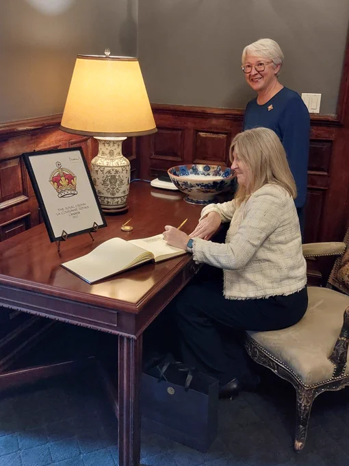 Alison Johnstone (seated) signing a visitor book, pictured with The Hon Edith Dumont, Lieutenant Governor of Ontario, in a wood-panelled room, with a polished desk and lit table lamp.