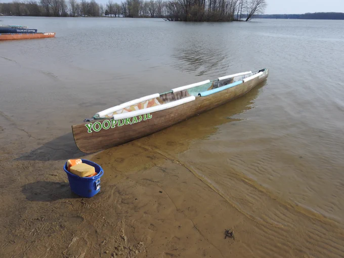 The concrete canoe “Yoopdrasil” rests partially in the water along a sandy shoreline, with a bucket of sponges nearby and trees lining the lake in the background.