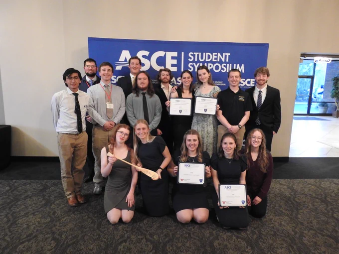 Concrete Canoe team pose in front of an ASCE Student Symposium backdrop, smiling and holding award certificates and a paddle to celebrate their competition results.