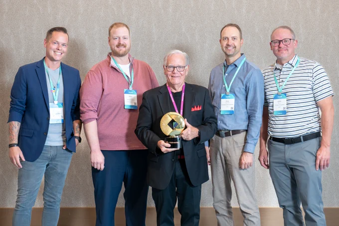 Five people standing together indoors, smiling. Jim Marous in the center holds a trophy with a prominent star design. They are all wearing name tags and conference lanyards. Neutral background.