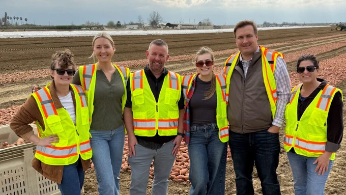 A group of six people wearing reflective safety vests stand together in a field. The background shows a tractor and expansive farmland under a cloudy sky.