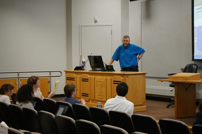 A lecturer stands at a podium at the front of the lecture hall. He is listening to a student ask a question.