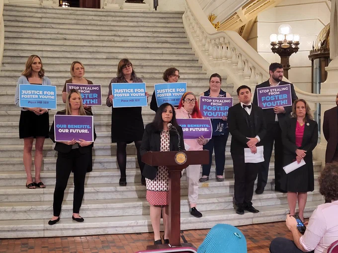 Photo of Director of Public Policy Dale Verchick speaking at a presser regarding house bill 151 in the Harrisburg Capitol building. Standing behind her are a group of individuals holding signs saying, "Justice for foster youth."