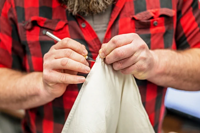 A person in a red and black plaid shirt skillfully using a seam ripper to remove stitches from a piece of off-white fabric.