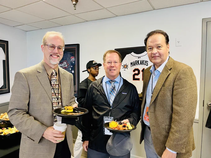 Three people stand together in an indoor setting, each holding food at groundbreaking ceremony for Harry Grove Stadium. Behind them are baseball jerseys displayed on the wall, including one with the name "Markakis" and the number 21.