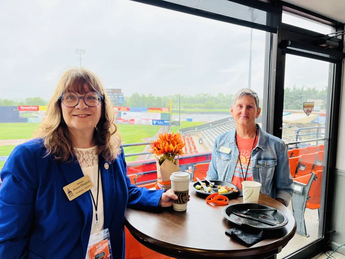 Two people are seated at a round table by a window overlooking a Harry Grove Stadium. One is wearing a blue jacket and name tag, while holding a coffee cup. The other is in a denim jacket, sitting beside a meal. Bright flowers decorate the table. Stadium details, including "Traveler" signs, are visible outside.