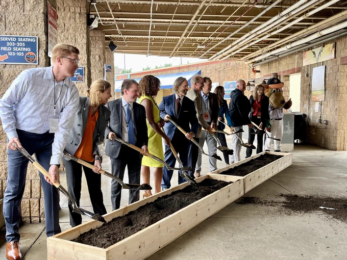 A group of individuals, including a character mascot, are participating in a ceremonial groundbreaking event inside a the Harry Grove Stadium concourse. They are holding shovels and standing over wooden boxes filled with soil. Signs for reserved seating sections are visible on the walls.