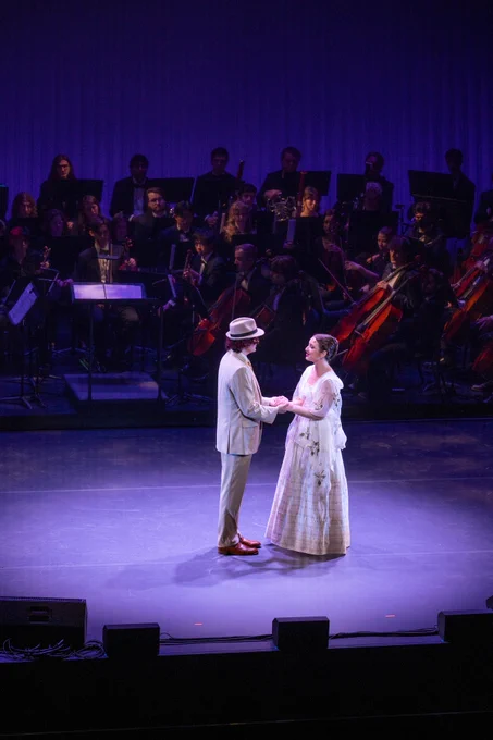 Two students wearing old fashioned costumes hold hands while singing on a stage. There is an orchestra behind them 