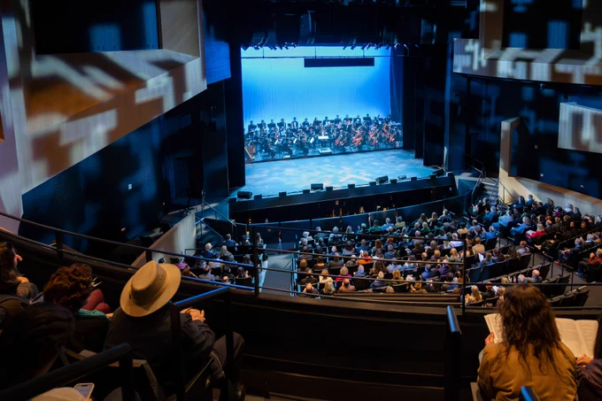 Audience sitting in seats inside a theater while an orchestra plays on stage